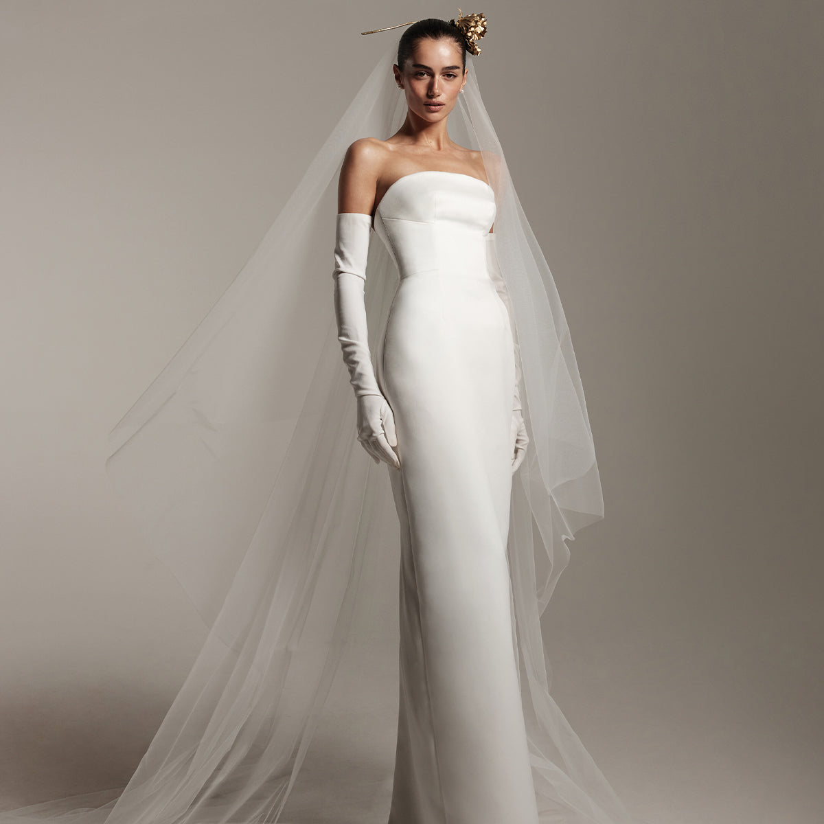 Model wearing Monique Lhuillier Spring 2026 Bridal white matte satin strapless Chandler column gown, white stretch crepe opera-length Chandler gloves, and two-tier tulle long veil with gold flower in hair