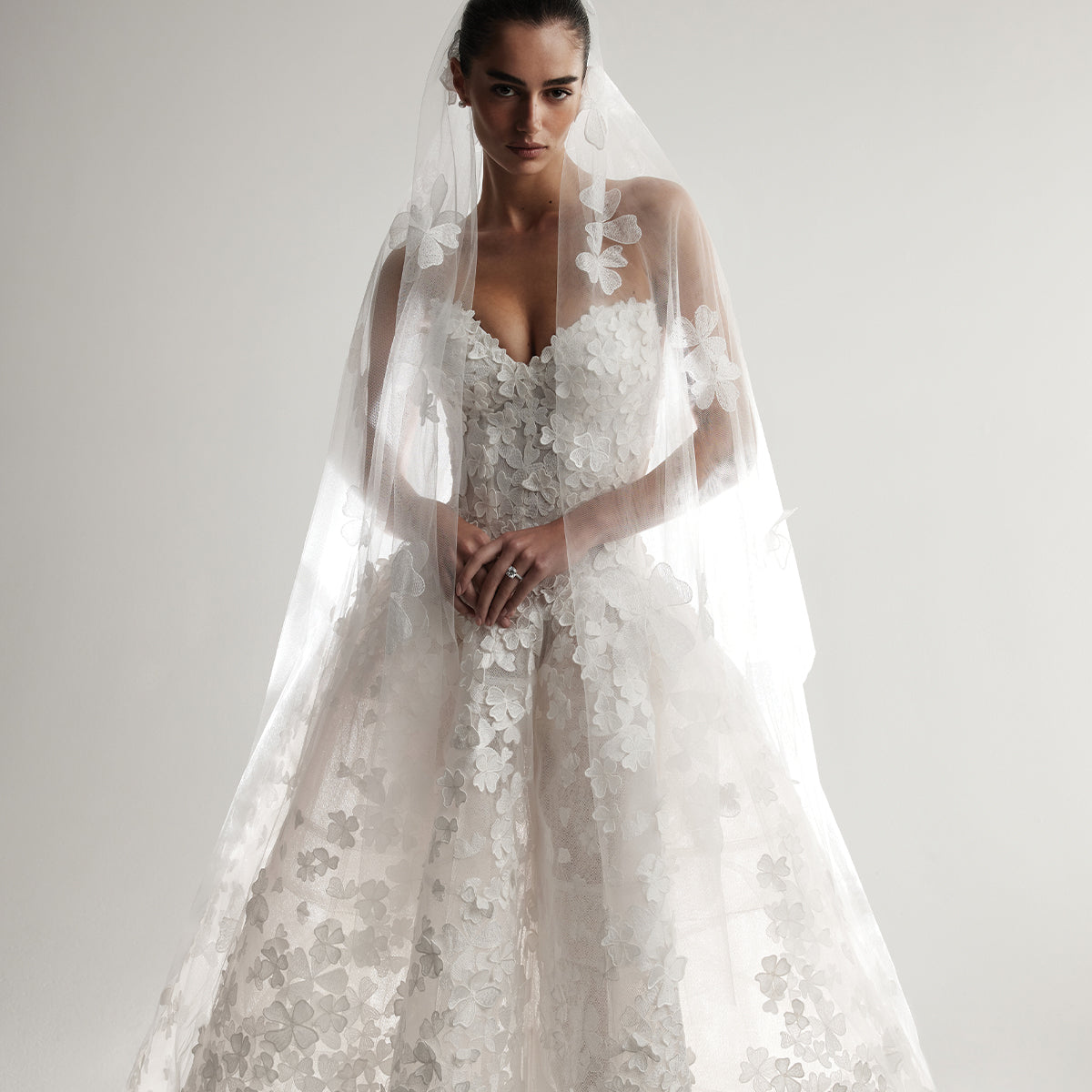 Model wearing Monique Lhuillier Spring 2026 Bridal white 3D embroidered tulle strapless elongated drop waist Clover ball gown with structured cone skirt and coordinating two tier Clover veil with floral embroidery