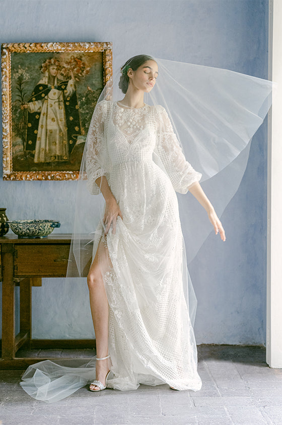 Woman wearing Monique Lhuillier Fall 2022 long sleeve white beaded sheath Emma gown with sheer veil over face