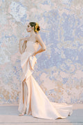 Woman wearing Monique Lhuillier Fall 2022 blush strapless draped trumpet Maxima gown with peplum and slit in skirt
