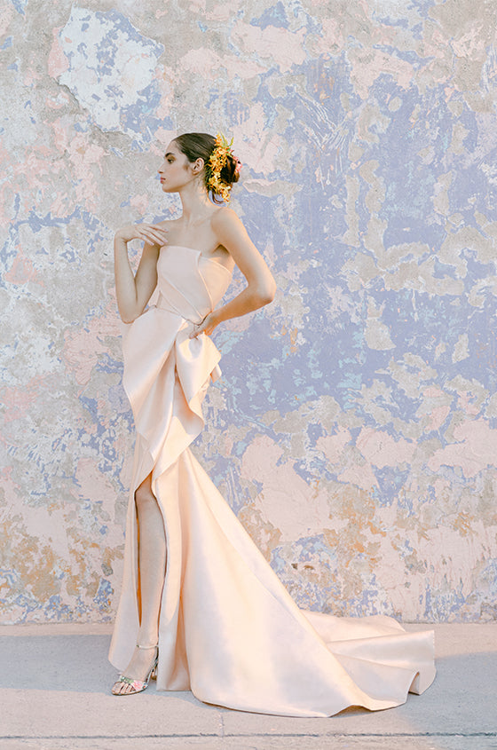 Woman wearing Monique Lhuillier Fall 2022 blush strapless draped trumpet Maxima gown with peplum and slit in skirt