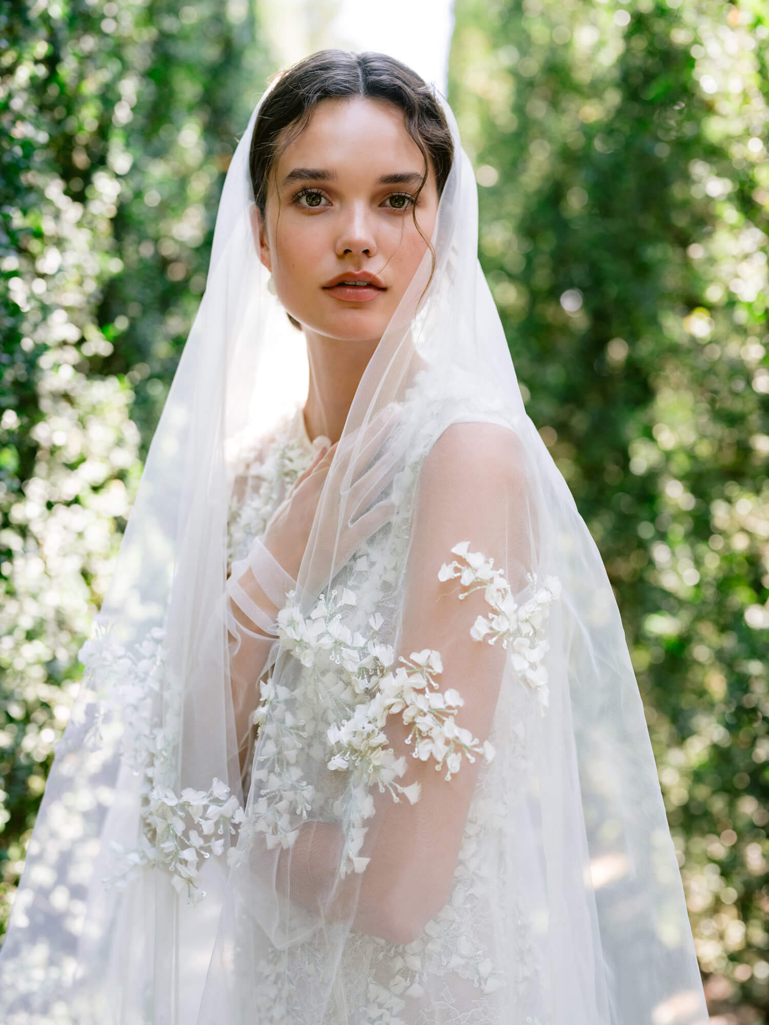 Woman wearing Monique Lhuillier Fall 2024 white fitted sleeveless 3-d embroidered Gabrielle gown with matching veil and tulle gloves