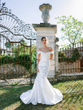 Woman wearing Monique Lhuillier Fall 2024 white satin Allure off-the-shoulder trumpet gown with draped neckline and skirt