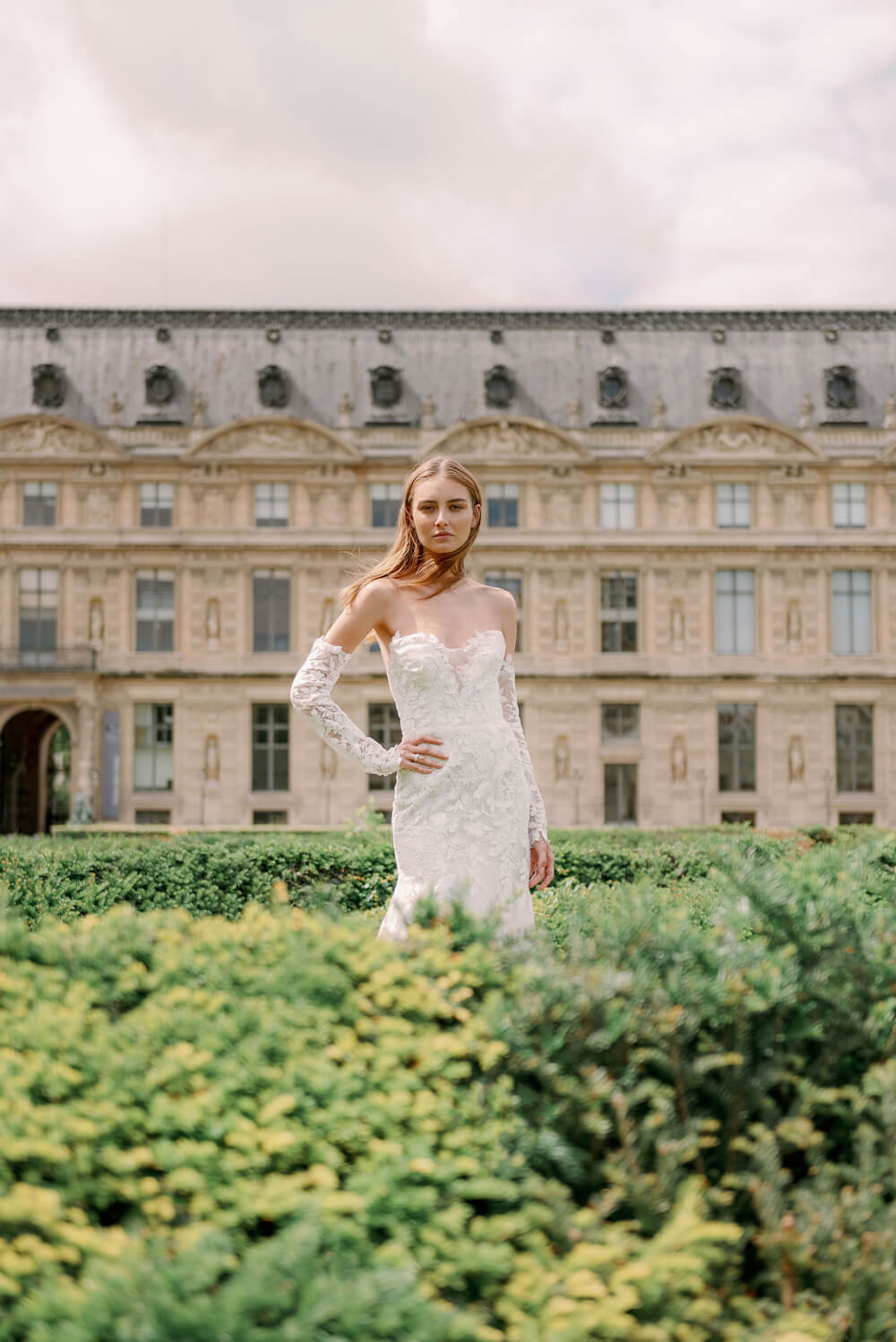 Woman wearing Monique Lhuillier Spring 2024 Bliss white embroidered lace BL24013 trumpet gown with detachable off-the-shoulder lace sleeves