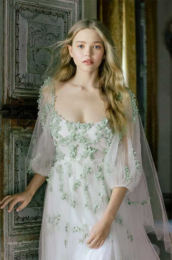 Woman wearing Spring 2022 Monique Lhuillier white 3/4 sleeve Wildflower sheath gown with green 3D floral embroidery