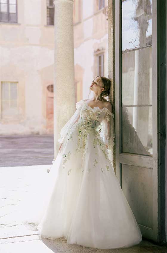 Woman wearing white off-the-shoulder Monique Lhuillier Spring 2022 Freesia a-line gown with billow sleeve and green 3D floral embroidery