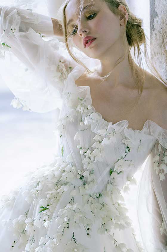 Woman wearing white off-the-shoulder Monique Lhuillier Spring 2022 Freesia a-line gown with billow sleeve and green 3D floral embroidery
