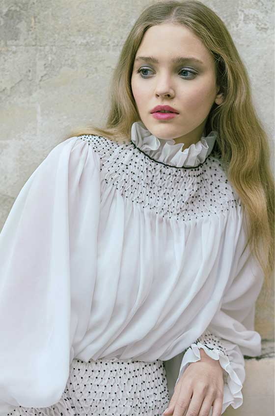 Woman wearing Monique Lhuillier Spring 2022 white high neck ruffle trim sheath gown with billow sleeve and black crystal detail style 22239