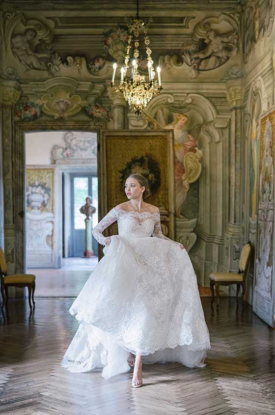 Woman wearing Monique Lhuillier Spring 2022 Fleur de Lis white lace a-line gown with matching off-the-shoulder long sleeve lace jacket