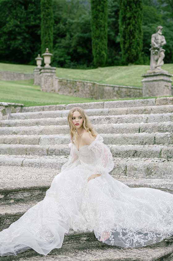 Woman wearing Monique Lhuillier Spring 2022 white embroidered a-line Splendid gown with off-the-shoulder billowed sleeve