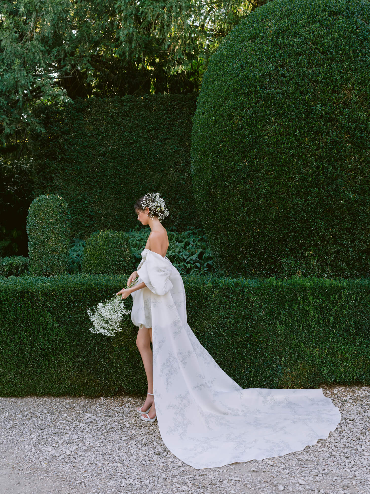 Woman wearing Monique Lhuillier Fall 2024 ivory printed gazar Avery bubble hem cocktail gown with detachable draped sleeve and cape train