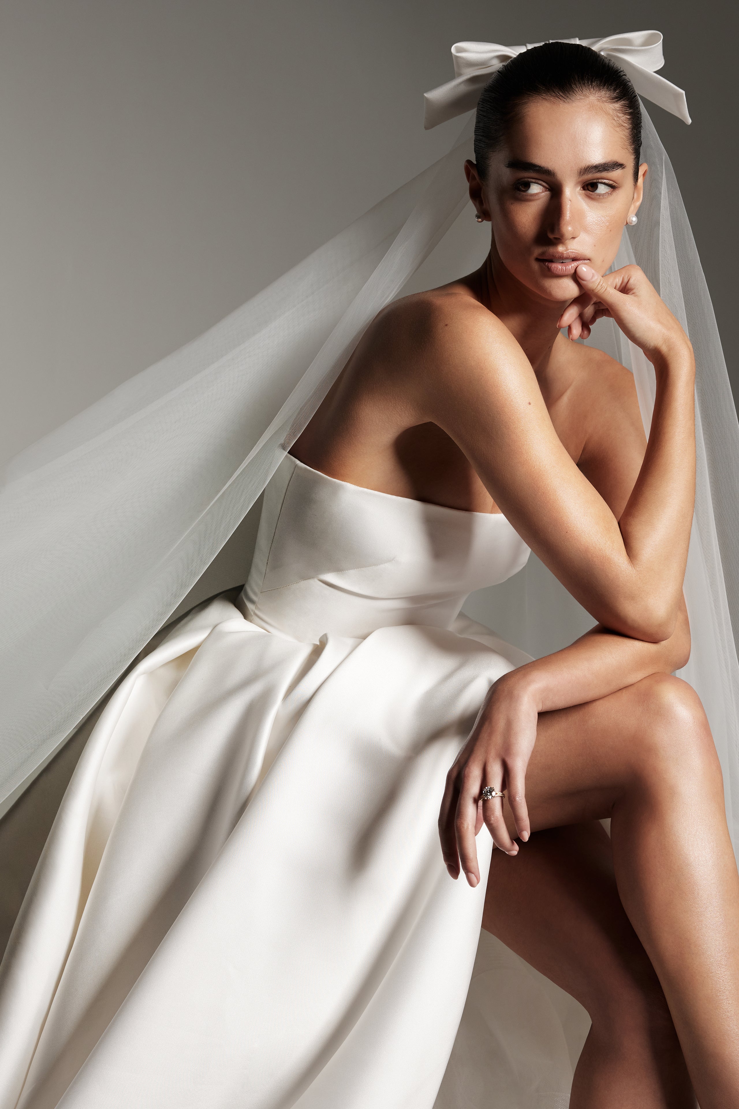 Model wearing Monique Lhuillier Spring 2026 Bridal white mikado Doheny strapless ball gown with slit and two tier Doheny veil with bow at comb.