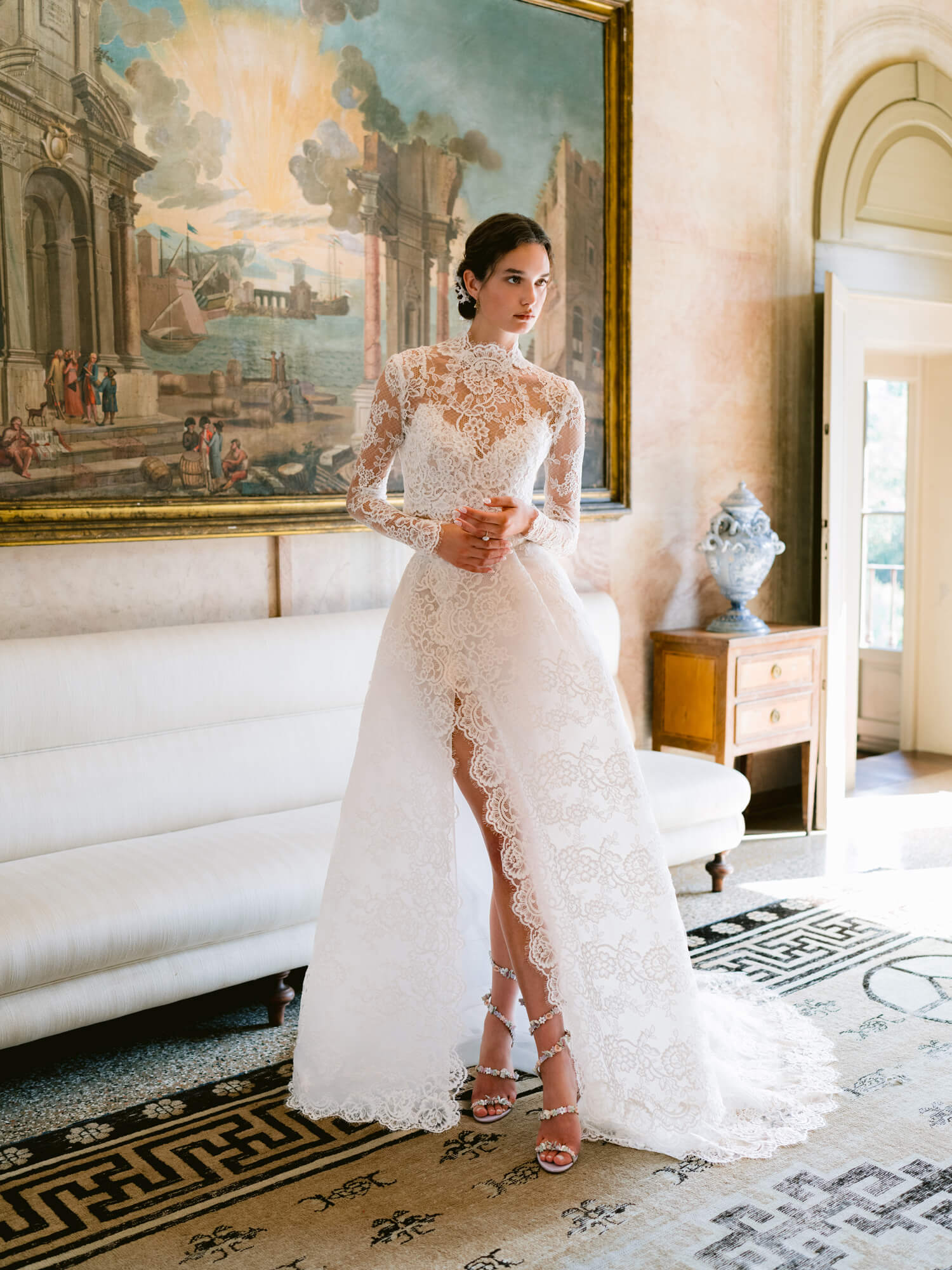 Woman wearing Monique Lhuillier Fall 2024 white Melrose lace bralette with lace long-sleeve high neck jacket and lace a-line skirt with slit
