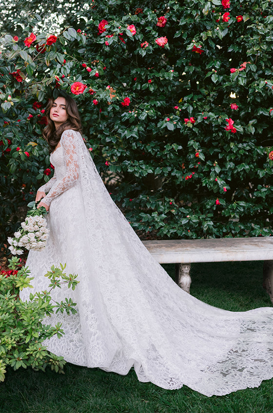 Woman wearing Monique Lhuillier Spring 2024 white lace scoop neck Kensington ballgown with matching long sleeve lace Kensington cape
