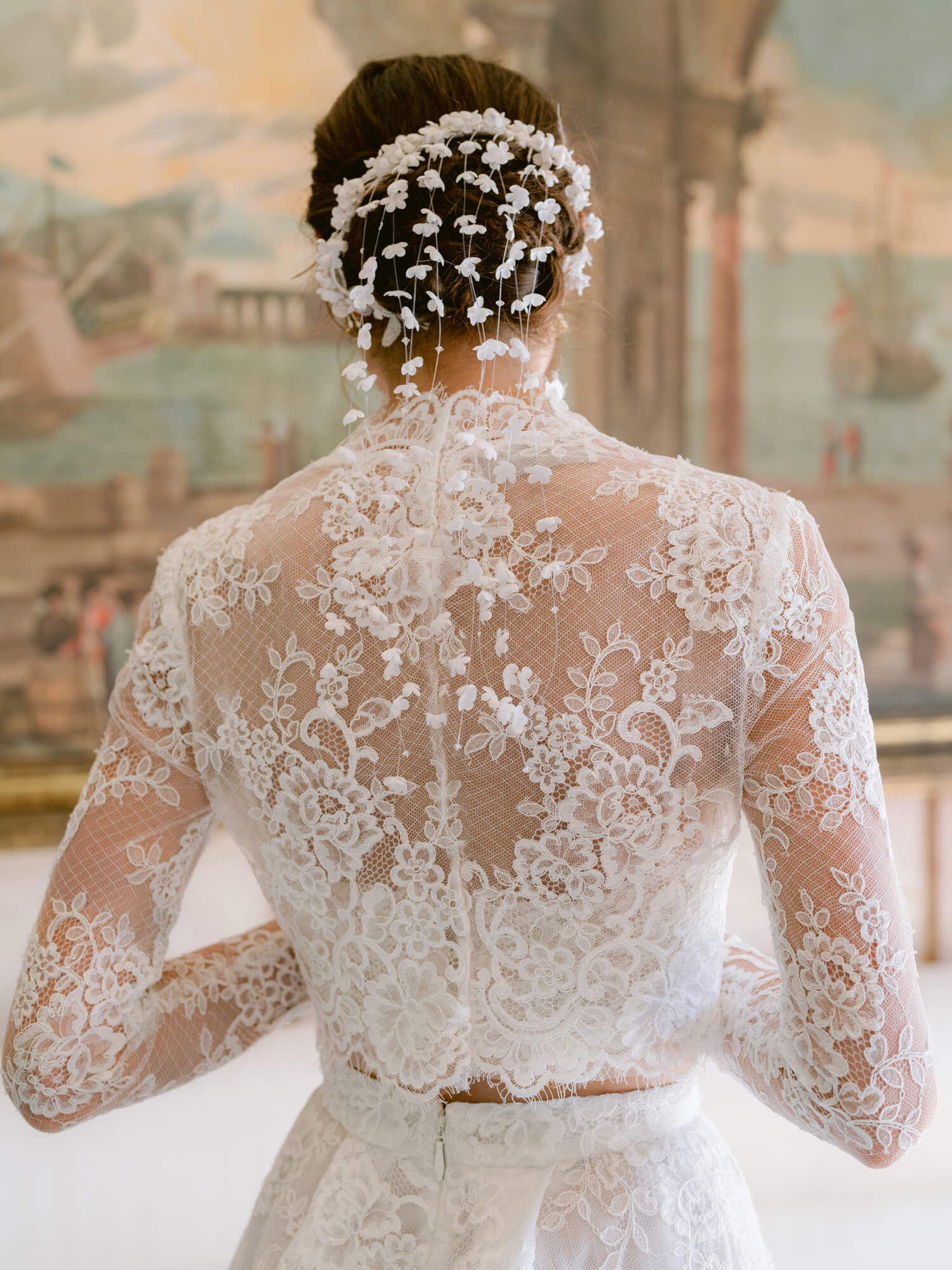 Woman wearing Monique Lhuillier Fall 2024 white Melrose lace bralette with lace long-sleeve high neck jacket and lace a-line skirt with slit