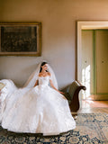 Woman wearing Monique Lhuillier Fall 2024 off-the-shoulder white lace Dolce Vita ballgown with lace embroidered veil