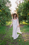 Woman wearing Monique Lhuillier Spring 2024 white strapless lace corseted Eden slip gown with matching long sleeve lace cape