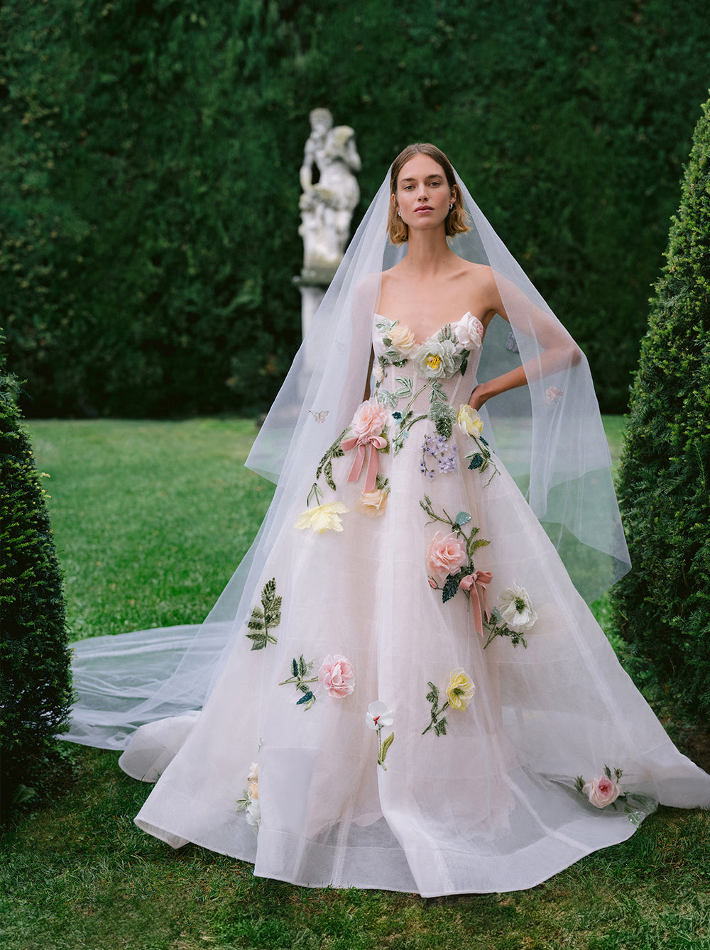 Monique Lhuillier Fall 2025 Bridal Amalfi Gown white and multi floral embroidered gown and Veil on model in garden.