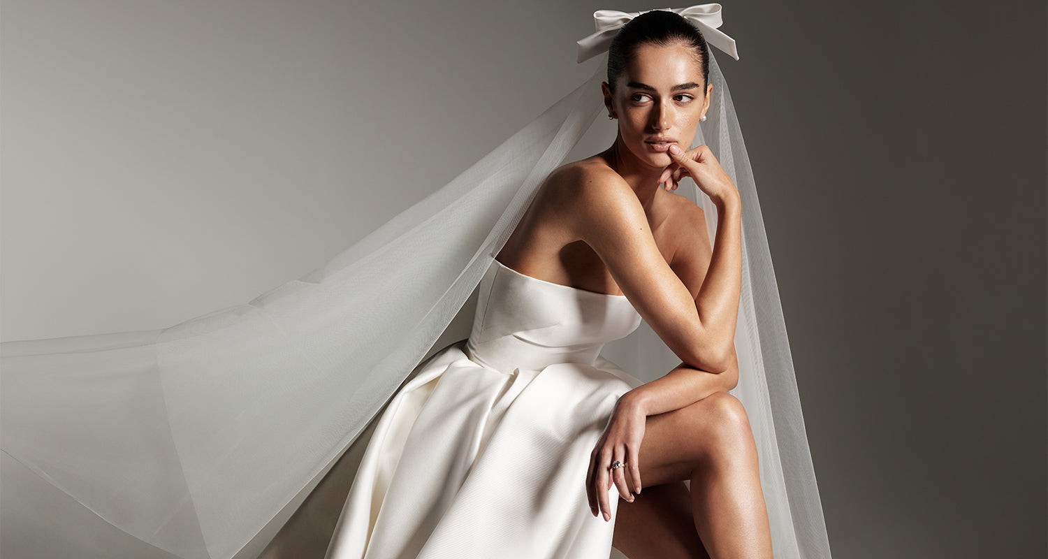 Model wearing Monique Lhuillier Spring 2026 Bridal white mikado Doheny strapless ball gown with slit and two tier Doheny veil with bow at comb.