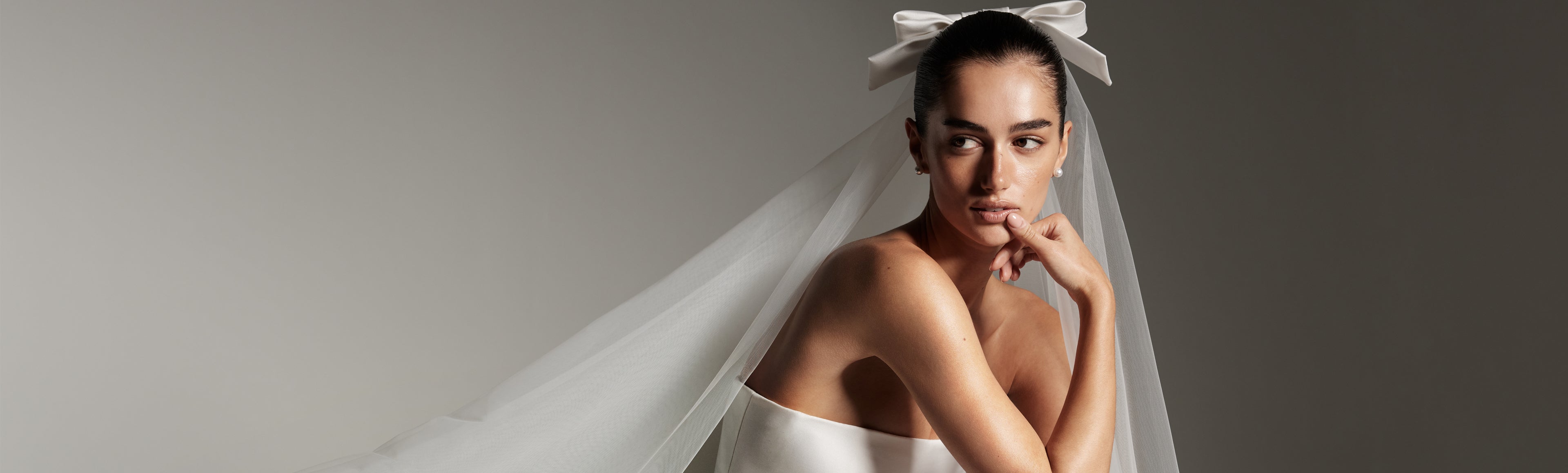 Model wearing Monique Lhuillier Spring 2026 Bridal white mikado Doheny strapless ball gown with slit and two tier Doheny veil with bow at comb.