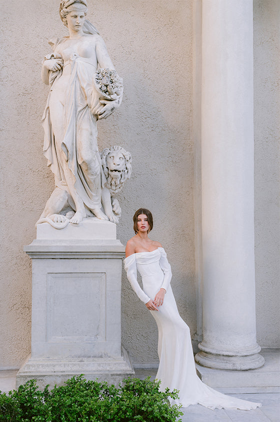 Woman wearing Monique Lhuillier Spring 2024 white off-the-shoulder long sleeve draped silk crepe Sussex column gown