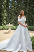 Woman wearing white Monique Lhuillier Spring 2021 duchess satin off-the-shoulder dropped waist a-line Marbella gown