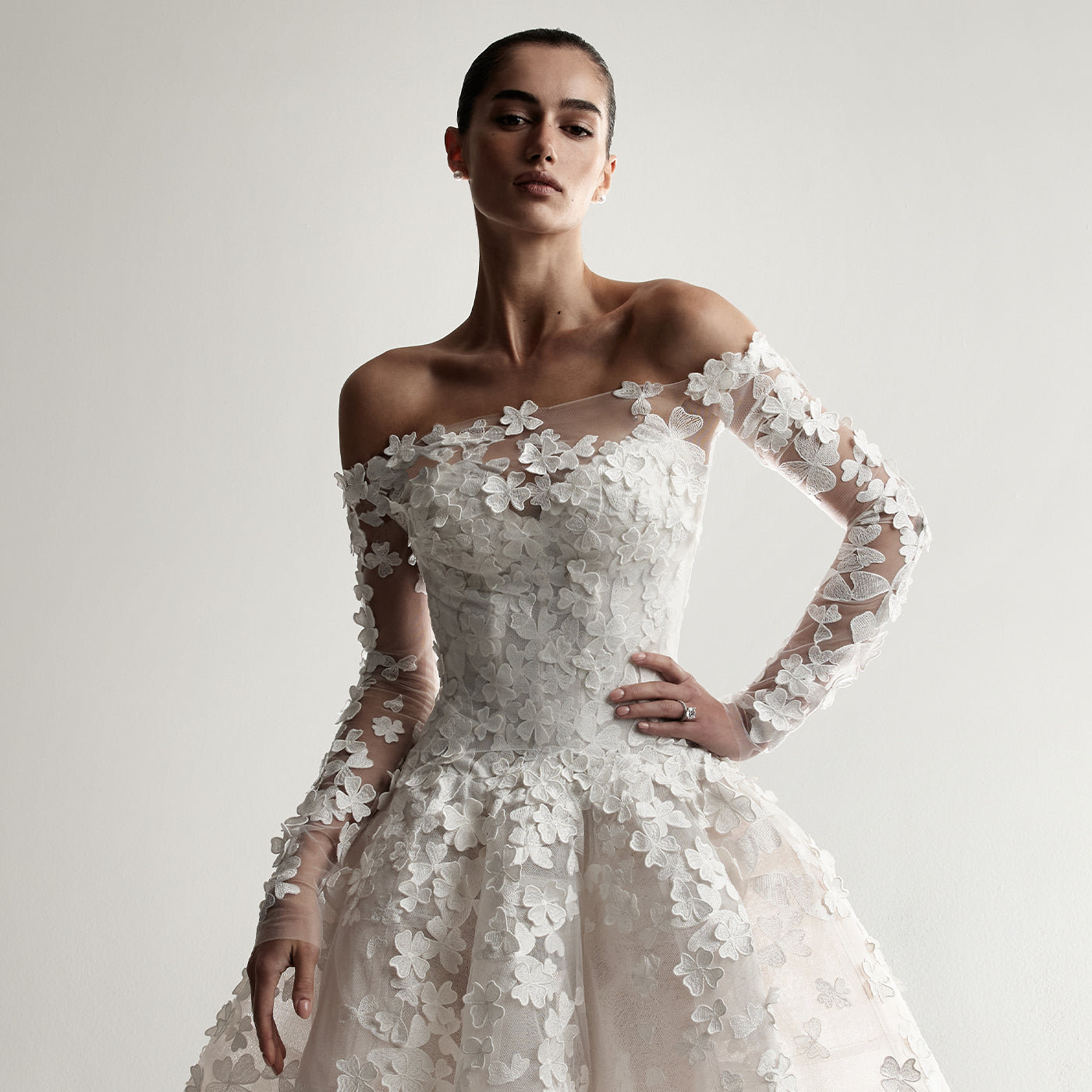 Model wearing Monique Lhuillier Spring 2026 Bridal white 3D embroidered tulle strapless elongated drop waist Clover ball gown with structured cone skirt and coordinating off-the-shoulder embroidered long sleeve Clover jacket 