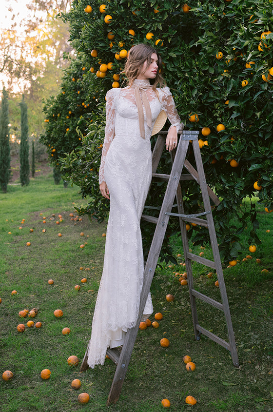 Woman wearing Monique Lhuillier Spring 2024 white lace high neck Victoria sheath gown with puff shoulder and long sleeves
