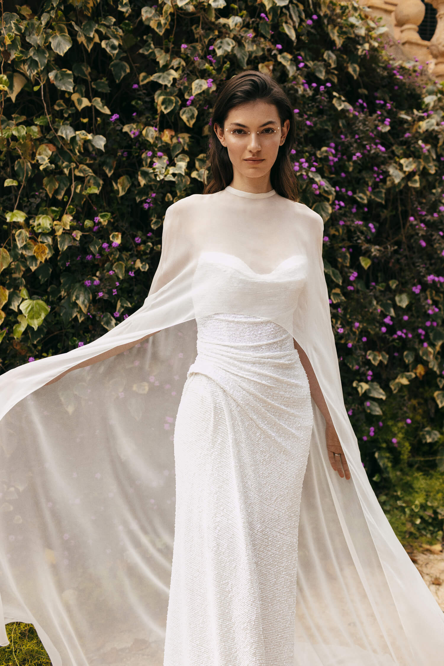 Woman wearing white sequin strapless Monique Lhuillier Spring 2023 Pearla draped column gown with white chiffon Pearla cape