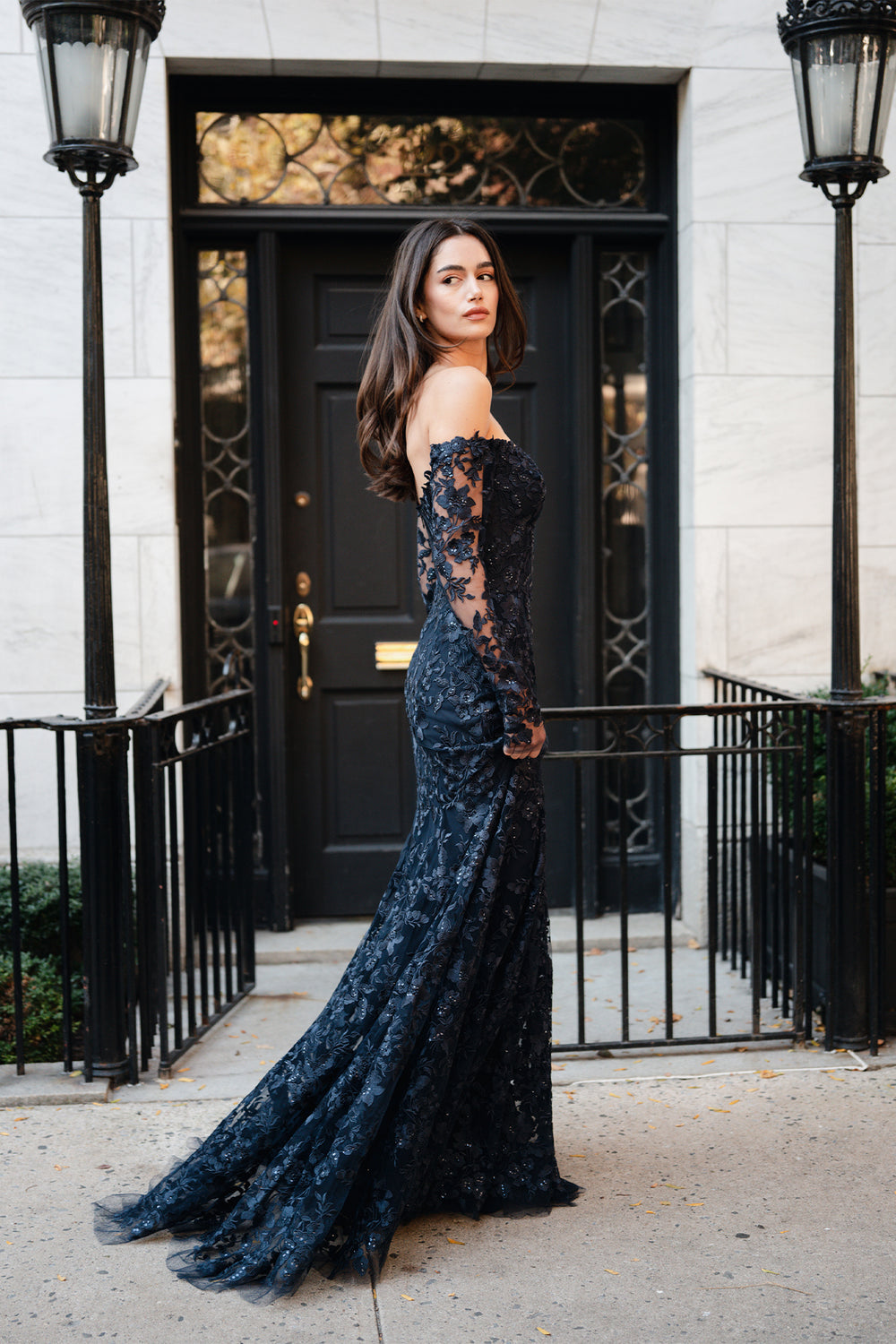 Monique Lhuillier Spring 2025 Off the shoulder, long sleeve trumpet gown with sweetheart neckline in Lapis/Noir re-embroidered tulle on model outside.