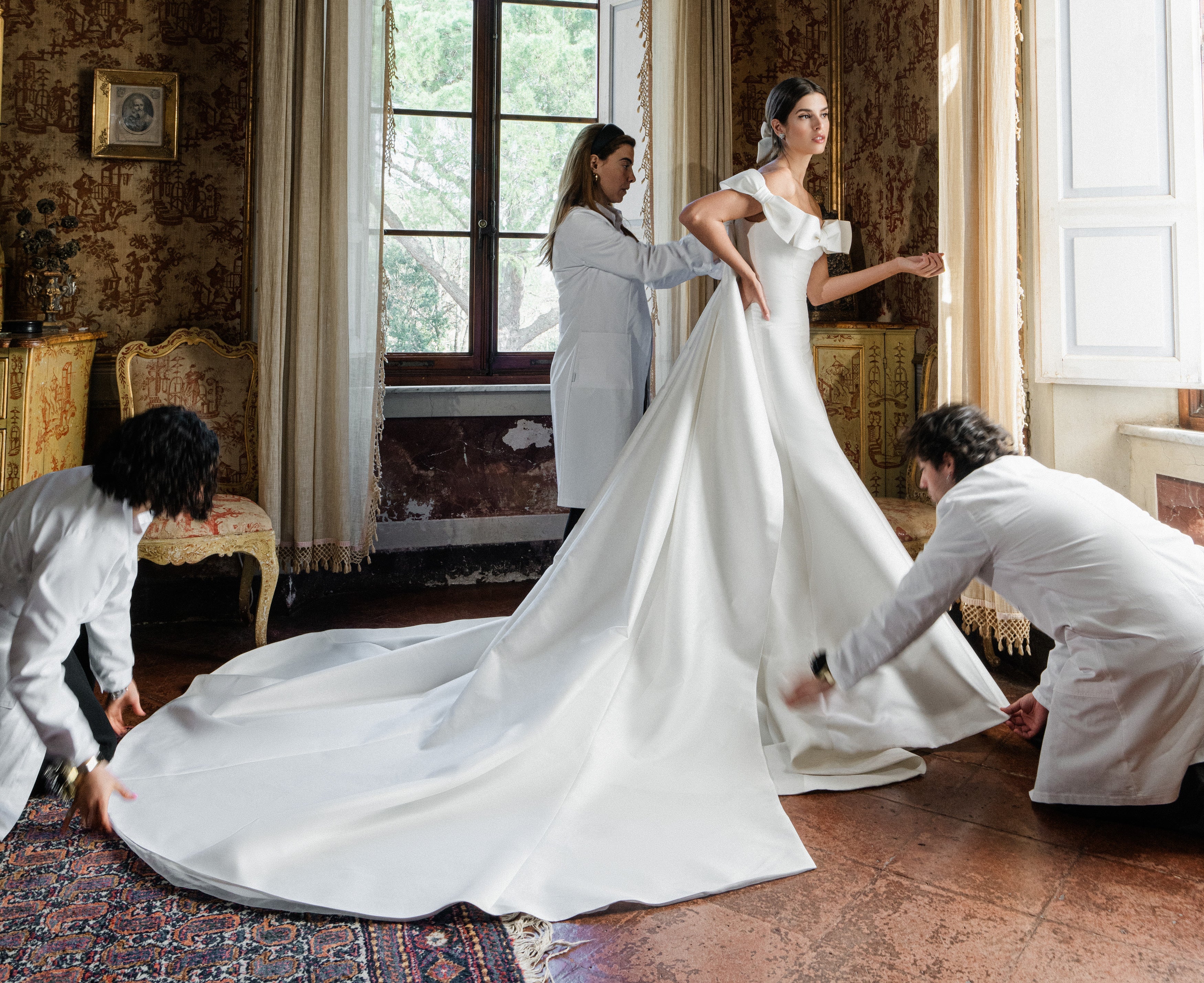 Monique Lhuillier Spring 2025 white mikado off-the-shoulder Mayfair trumpet gown with bow detail at neckline and train on model by window 
