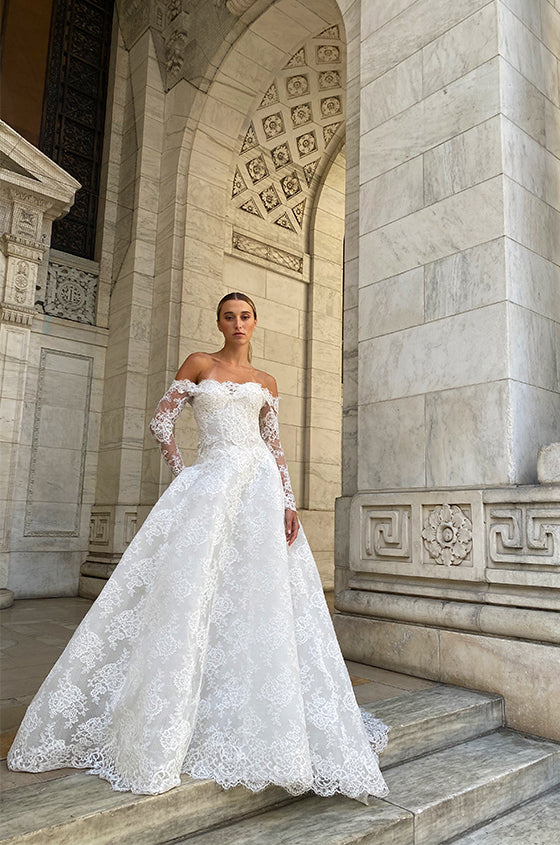 Woman wearing Monique Lhuillier Spring 2022 Fleur de Lis white lace a-line gown with matching off-the-shoulder long sleeve lace jacket