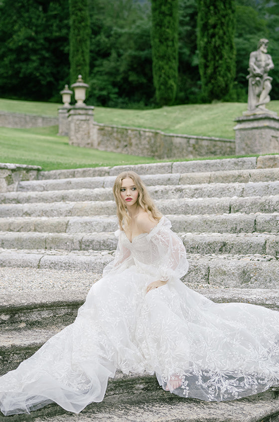 Woman wearing Monique Lhuillier Spring 2022 white embroidered a-line Splendid gown with off-the-shoulder billowed sleeve