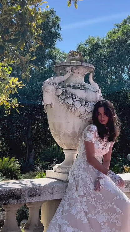 Women leaning on railing in Monique Lhuillier Bridal Spring 2024 blush and white floral embroidered Antoinette gown, jacket and gloves. 