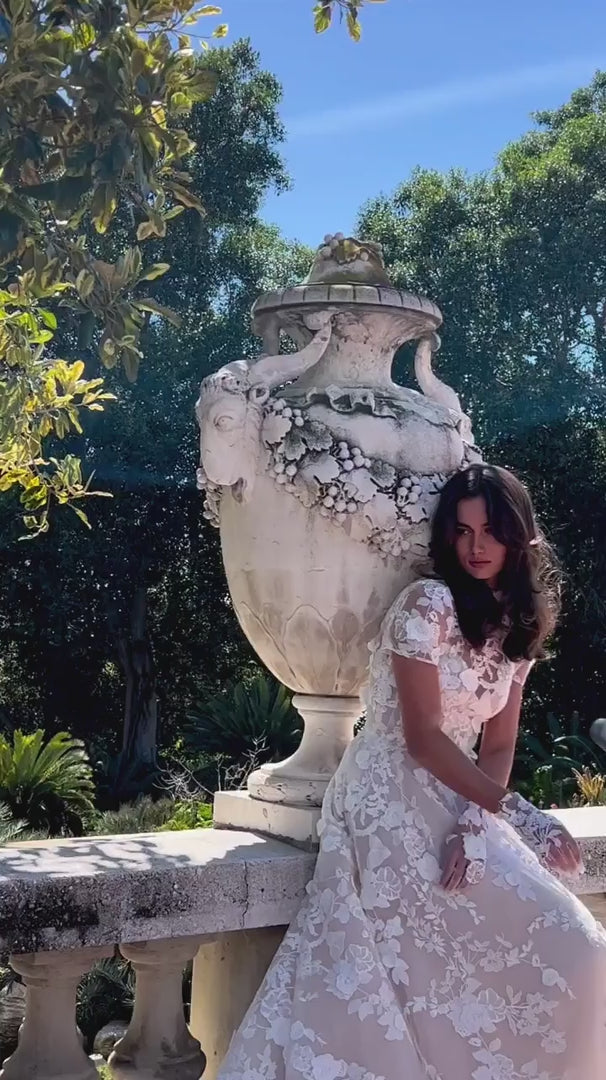 Women leaning on railing in Monique Lhuillier Bridal Spring 2024 blush and white floral embroidered Antoinette gown, jacket and gloves. 