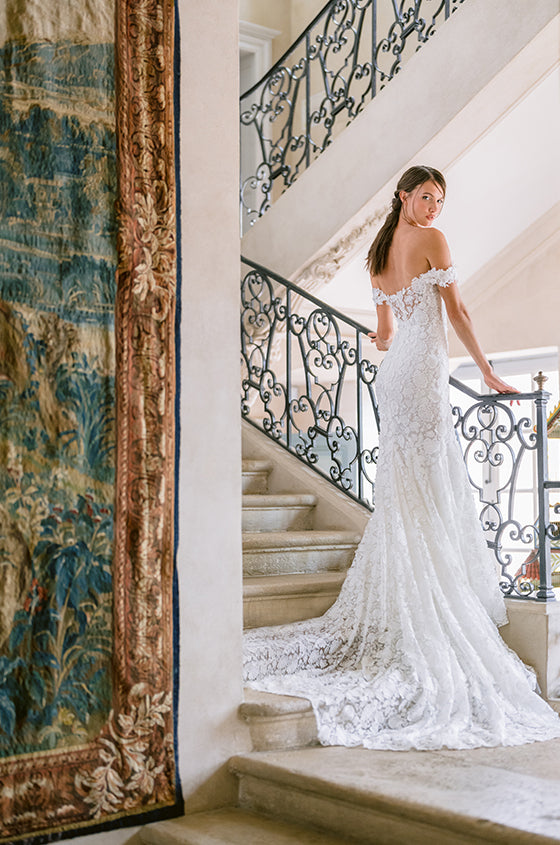 Woman wearing Monique Lhuillier Fall 2023 white off-the-shoulder fitted lace Desire trumpet gown with long train